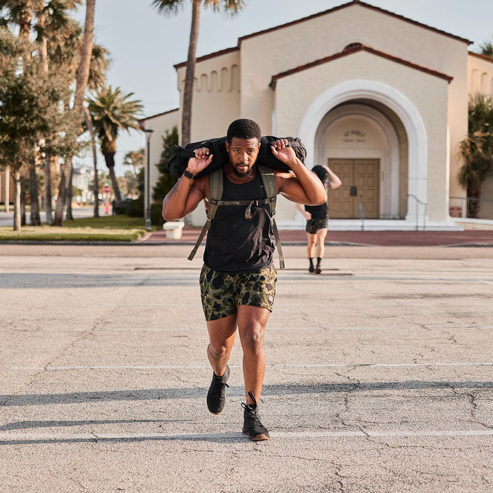 A man works out with the GORUCK Ballistic Trainers - Mid Top - Black + Gum featuring a Black Reflective Spearhead, known for their CORDURA® Ballistic Nylon construction that offers superior durability, near a building adorned with palm trees in the background.