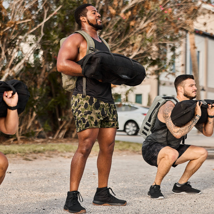 Men outdoors are performing squats with weighted backpacks during a workout, ensuring stability and enhanced strength. The Ballistic Trainers from GORUCK, featuring durable CORDURA® Ballistic Nylon material in a Mid Top design, offer robust support for each squat, making them both challenging and secure.