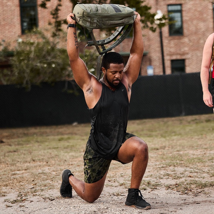 A man exercises outdoors, demonstrating balance as he lifts his GORUCK Ballistic Trainers - Mid Top - Black + Gum with Black Reflective Spearhead overhead while kneeling on one knee.