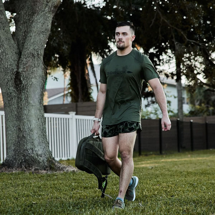 A man in a green GORUCK ToughMesh Men's Performance Tee and camouflage shorts walks on grass, holding a backpack in one hand. He is outdoors in a park-like setting, with trees and a fence visible in the background.