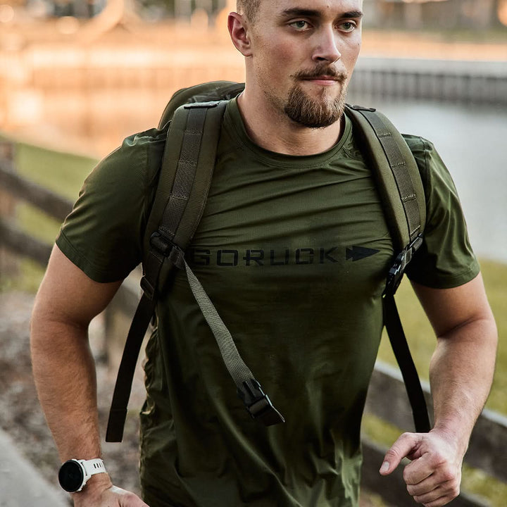 A man in a green GORUCK Men’s Performance Tee - ToughMesh and watch walks with a backpack near a wooden fence and a body of water. The early morning or late afternoon light creates a warm glow, highlighting his determined expression as the quick-drying performance fabric keeps him comfortable.