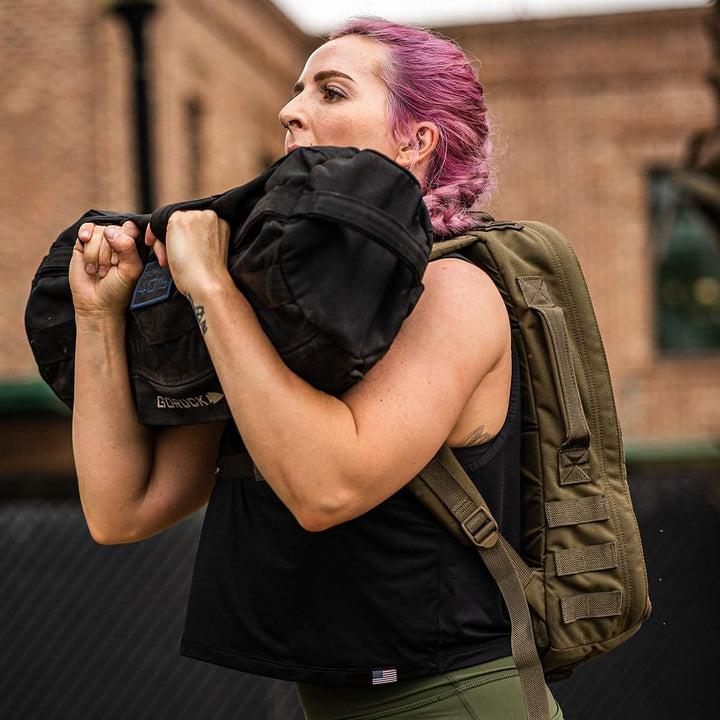 A person with pink hair is outdoors, lifting a heavy sandbag while wearing GORUCK's black Women’s Performance Tank - ToughMesh and green pants, accompanied by a backpack. A brick building and a fence in the background add to the rugged charm reminiscent of the Italian Alps.