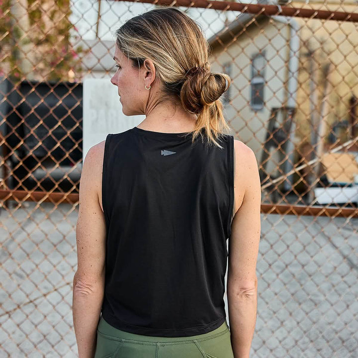 A woman with brown hair styled in a bun stands outside, her back facing the camera. She is wearing olive green pants and the Women's Performance Tank - ToughMesh by GORUCK. A chain-link fence and distant buildings extend toward the horizon, creating a scene reminiscent of the Italian Alps.