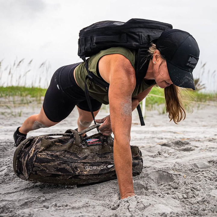 A person in a black cap and the Women’s Performance Tank - ToughMesh by GORUCK is doing a push-up on a sandy beach, grasping a camouflage-patterned bag with one hand. The sky is overcast, and grass can be seen in the background.