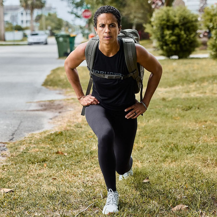 A person in a GORUCK Women’s Performance Tank - ToughMesh, leggings, and sneakers performs lunges on a grassy area. They have a backpack on and are exercising outdoors on a suburban street with bushes and trees visible in the background.