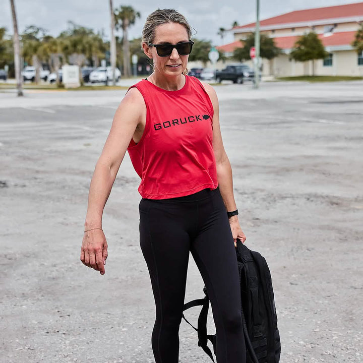 A woman in a red GORUCK Women’s Performance Tank made from ToughMesh fabric and black leggings is seen walking outdoors. She carries a black bag, wears sunglasses and a watch, with a backdrop of a street lined with buildings. The sky is overcast.