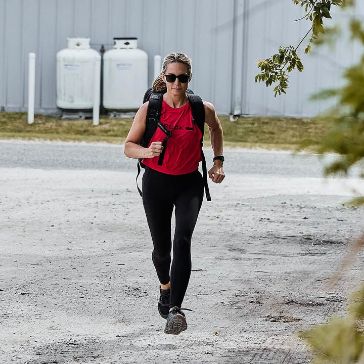 Wearing a Women’s Performance Tank - ToughMesh from GORUCK, black leggings, and sunglasses, a person jogs briskly along a gravel path with a backpack. With greenery and propane tanks in the background, they move with precision as if training for a trek through the Italian Alps.