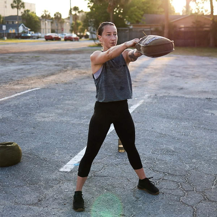 A person exercises outdoors, holding a weighted sandbag at shoulder height. They wear the GORUCK Women’s Performance Tank made from ToughMesh fabric paired with black leggings. The sun is setting, casting a warm glow across the pavement and nearby trees reminiscent of the Italian Alps' serene beauty.