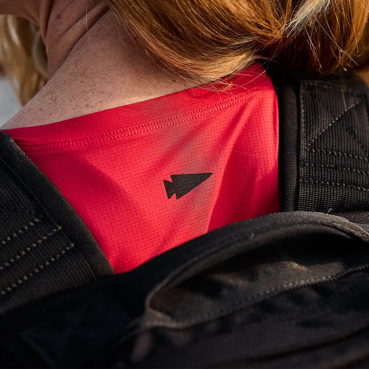 Close-up of a person wearing the bright red GORUCK Women’s Performance Tank - ToughMesh featuring a black arrowhead symbol on the back, partially obscured by black backpack straps. The person's shoulder and a hint of hair are visible, suggesting they're prepared to tackle the trails in the Italian Alps.