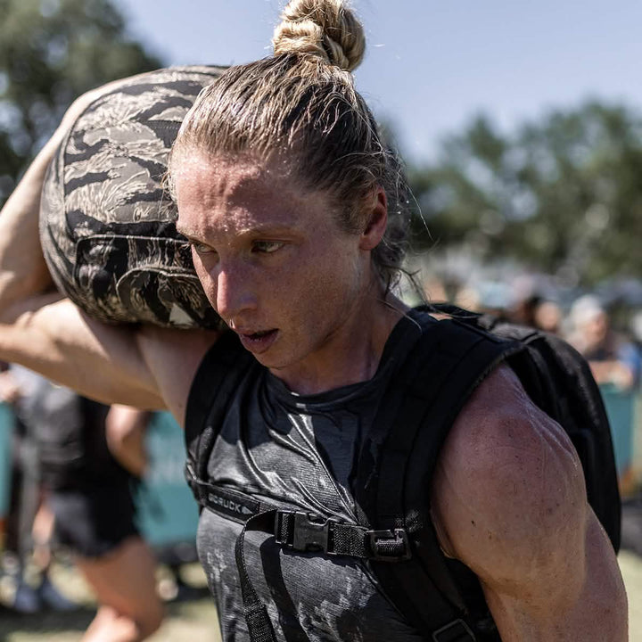 A determined athlete with a topknot hoists a hefty sandbag onto their shoulder during an outdoor competition. Wearing the Women's Performance Tank by GORUCK, crafted with ToughMesh™ fabric, they push through the event, as blurred spectators and trees surround them, evoking the rugged landscapes of the Italian Alps.