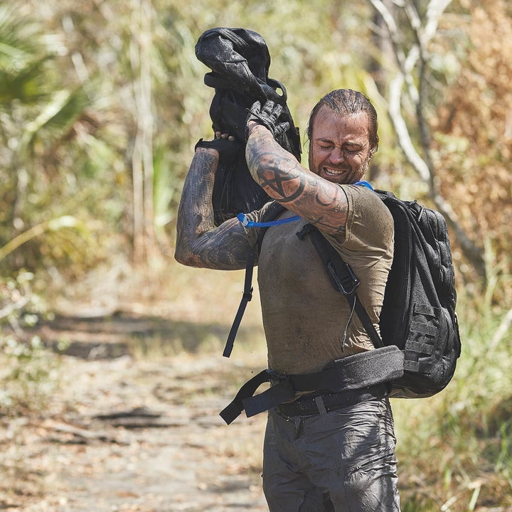 A man with tattoos adjusts his GORUCK backpack, equipped with a padded hip belt, on a dirt path in a wooded area.