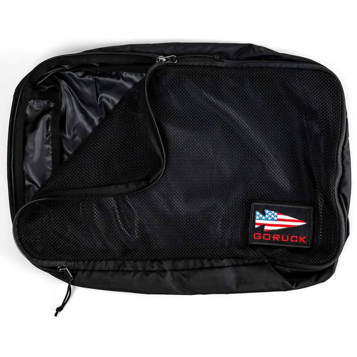 A GORUCK Packing Cube in black, made from ultra-light nylon and featuring a mesh cover, is partially unzipped to show its inner compartment. It proudly displays an American flag patch and the GORUCK logo in the bottom right corner, making it a perfect travel companion.