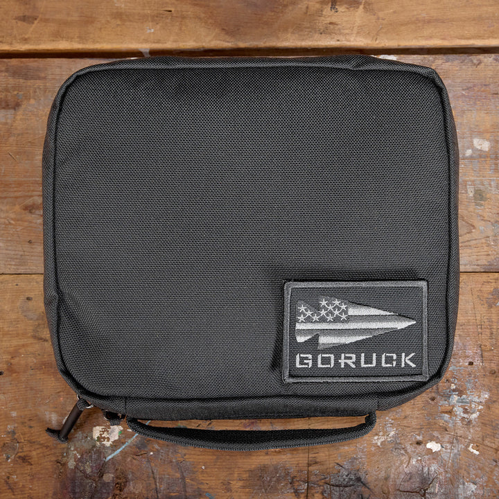 The GR2 Field Pocket, a rugged black zippered pouch featuring a "GORUCK" patch, is crafted from durable 1000D Cordura and rests on a worn wooden surface. It's the perfect electronics organizer for your adventures.