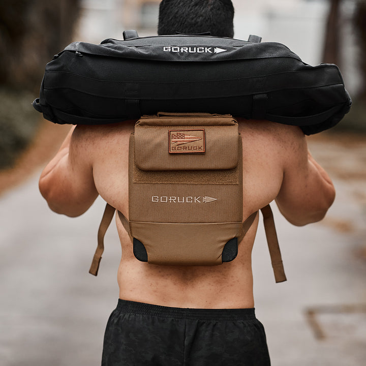 A person carries a black GORUCK Ruck Plate Carrier 3.0 over their shoulders and wears a tan GORUCK backpack with ergonomic lumbar support, both crafted from durable ballistic nylon, as they walk along an outdoor path.