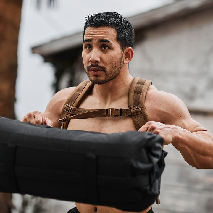 In an outdoor setting, someone is lifting a sandbag while wearing the GORUCK Ruck Plate Carrier 3.0, a tan harness made from durable ballistic nylon with ergonomic lumbar support, all while maintaining a focused expression.