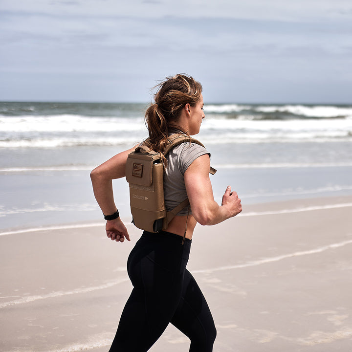 A woman jogs along the beach with a GORUCK Ruck Plate Carrier 3.0, made of durable ballistic nylon, while ocean waves crash in the background.