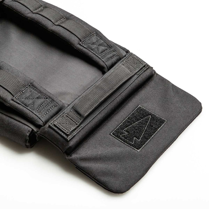 A close-up of the GORUCK Ruck Plate Carrier 3.0, a black tactical bag made from durable 1000D CORDURA, showcases its black Velcro flap adorned with a patch featuring a stylized arrowhead symbol. The bag is designed with reinforced stitching, ergonomic lumbar support, and padded straps against a plain white background.