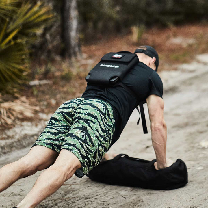 Wearing a backward cap and camo shorts, a person executes a plank exercise on a dirt path, adding an extra challenge with the GORUCK Ruck Plate Carrier 3.0, amidst a backdrop of trees.