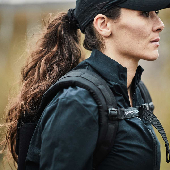 A woman with long, wavy hair styled in a ponytail is wearing a black cap and jacket. Outdoors, with blurred greenery as the backdrop, she carries a durable GORUCK Ruck Plate Carrier 3.0 backpack made of 1000D CORDURA. Her expression is focused as she gazes off to the side.