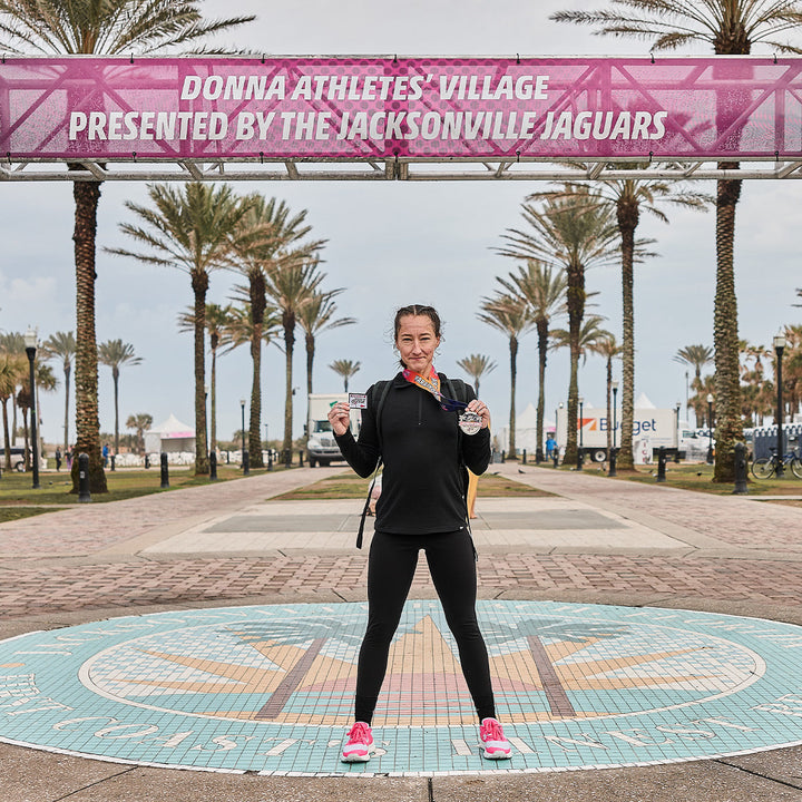 At Donna Athletes' Village, a woman beams with pride, a medal in one hand and a drink in the other. Her Rough Runner - Hot Pink by GORUCK, featuring an ultra-supportive design and versatile road-to-trail outsole, have clearly carried her to victory today.