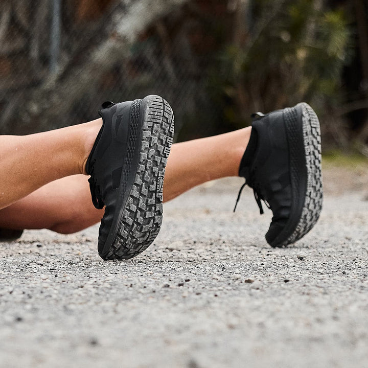 A person lies on the gravel wearing GORUCK's Rough Runner - Blackout shoes, with soles facing upward to reveal their versatile rugged road-to-trail outsole.