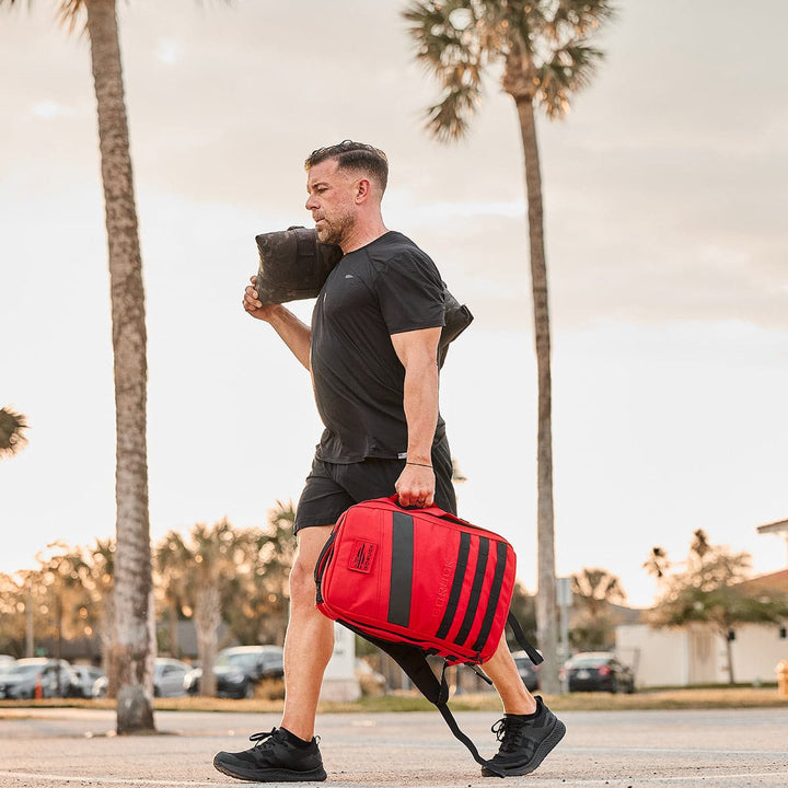 A person with a red backpack and water bottle strides confidently, their Rough Runner - Blackout shoes from GORUCK softly crunching on the path. Surrounded by palm trees, they enjoy the support of a Gradient Density EVA midsole and road-to-trail outsole as they explore the tropical paradise.