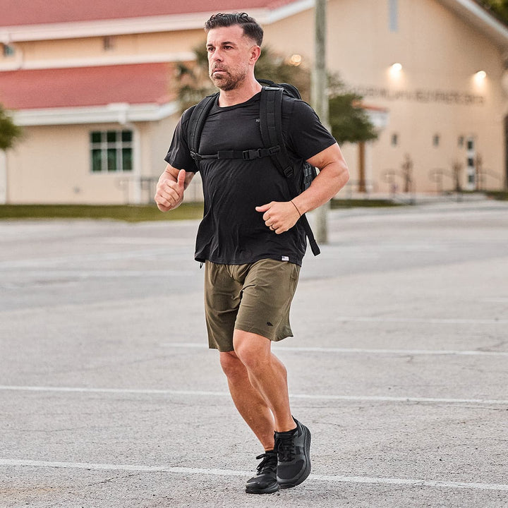 A man jogs through the parking lot near a building, his backpack bouncing gently with each step. His Rough Runner - Blackout shoes from GORUCK, equipped with a Gradient Density EVA midsole and road-to-trail outsole, provide the perfect blend of comfort and adaptability for any terrain.