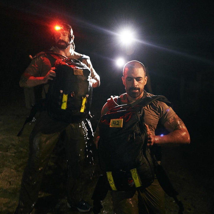 Two men in wet gear carry backpacks outfitted with GORUCK's Reflective Ruck Bands, their path lit by headlamps to enhance nighttime visibility.