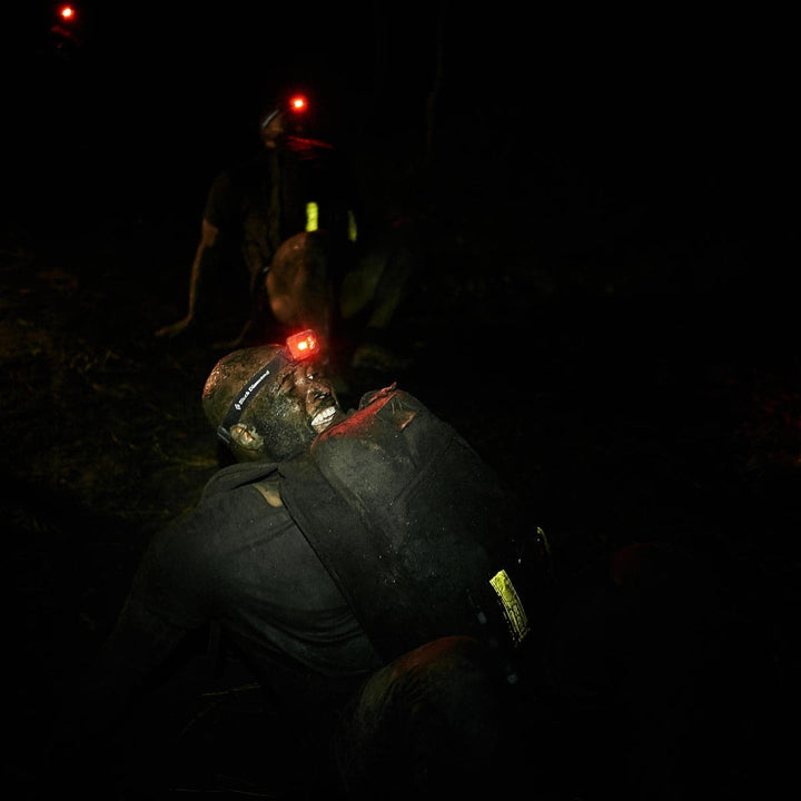 A person with a headlamp, covered in mud, crawls through the night during a challenge, their GORUCK Reflective Ruck Bands ensuring nighttime visibility.