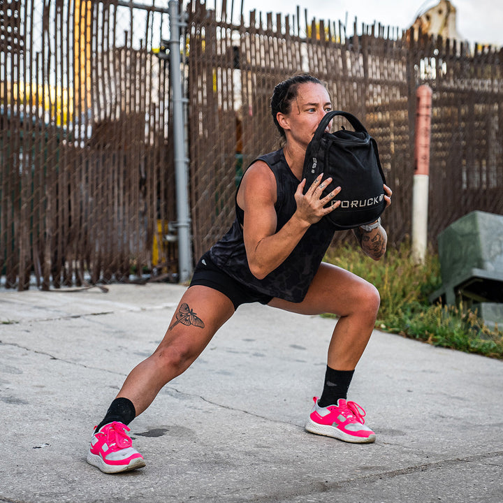 While exercising outdoors, this individual performs side lunges with a weighted bag, dressed in a black outfit and GORUCK's Rough Runner sneakers in Hot Pink, which feature a versatile road-to-trail outsole.