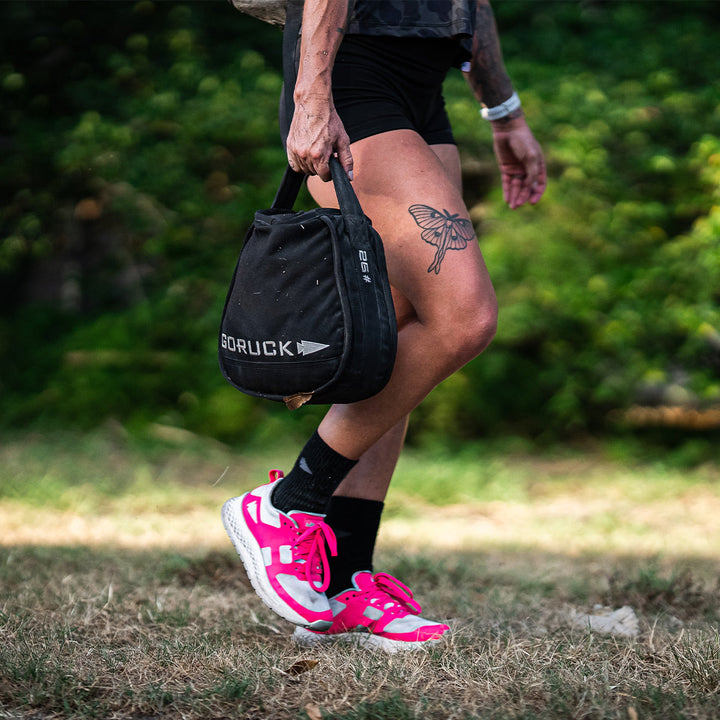 A person walks on grass, wearing the ultra-supportive Rough Runner - Hot Pink shoes from GORUCK, which feature a versatile road-to-trail outsole and GRADIENT DENSITY™ EVA MIDSOLE for comfort and stability. They're holding a GORUCK bag, with their bee tattoo visible on their thigh.