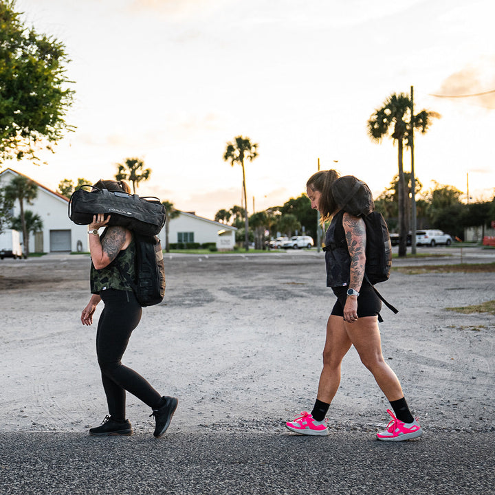 Two people walking on a road carrying large backpacks under a cloudy sky, their Rough Runner - Hot Pink shoes by GORUCK featuring an ultra-supportive running design with a versatile road-to-trail outsole ready for any terrain.