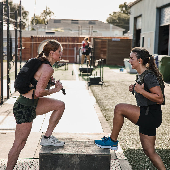 Two women engage in step-up exercises on a wooden box outdoors, clad in athletic attire and GORUCK Rucker 4.0 backpacks. They exchange smiles amidst gym equipment and a fenced area, all under a clear sky, seamlessly combining fitness with the thrill of rucking.