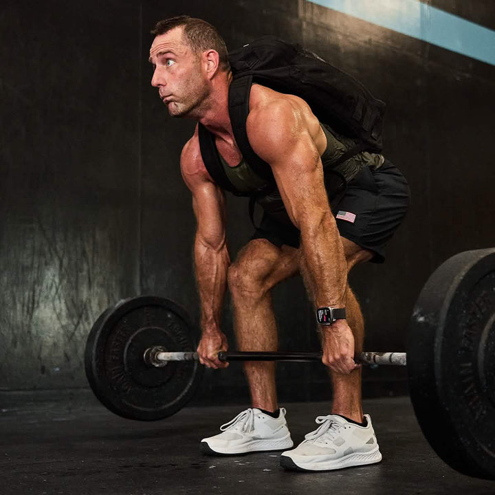 A man lifts a barbell at the gym, focused and experienced in rucking for endurance, wearing a black GORUCK Rucker 4.0 backpack and white sneakers.