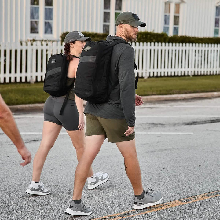 Two people, dressed in athletic gear, walk along a road past a white fence and building while wearing GORUCK Rucker 4.0 backpacks.