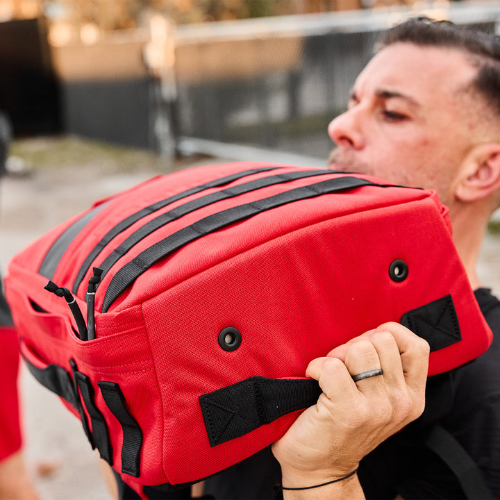 A person is outdoors, lifting a red sandbag with black straps, much like hefting the GORUCK Rucker 4.0 during an intense rucking session.