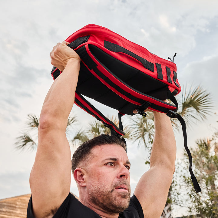 A man is rucking outdoors, lifting a red and black GORUCK Rucker 4.0 overhead with palm trees swaying gently in the background.