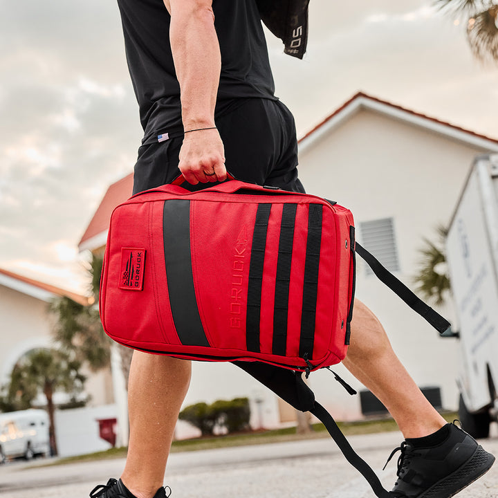 A person with a GORUCK Rucker 4.0 is walking outdoors, showcasing a vibrant red backpack against the backdrop of a charming house and lush trees.