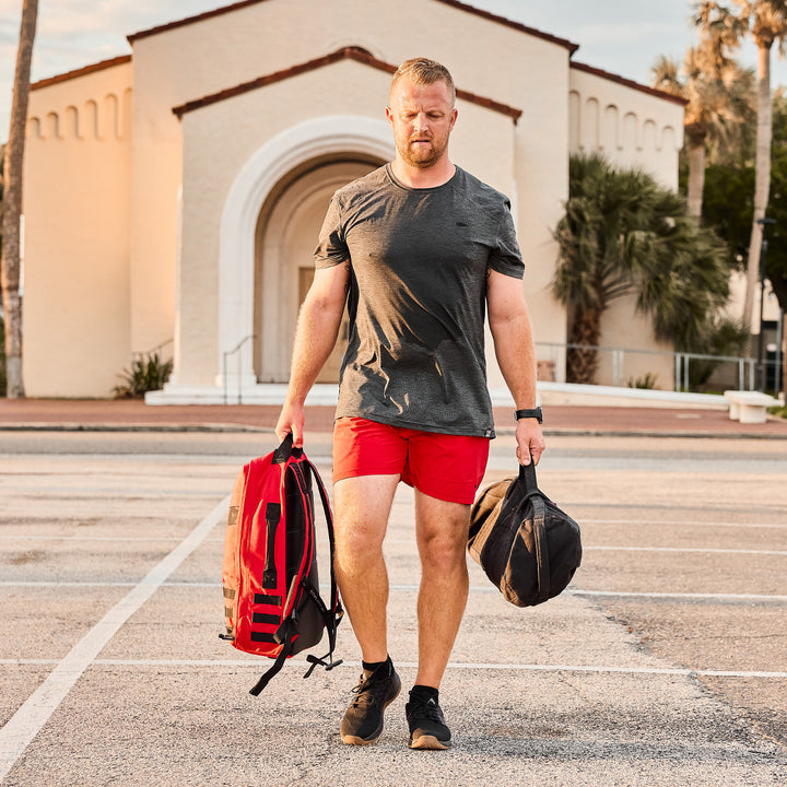 A man in red shorts and a gray GORUCK Rucker 4.0 shirt is rucking through the parking lot, effortlessly carrying bags. A building stands prominently in the background.