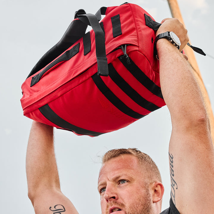 A person holding a red GORUCK Rucker 4.0 overhead, revealing arm tattoos and wearing a black watch.