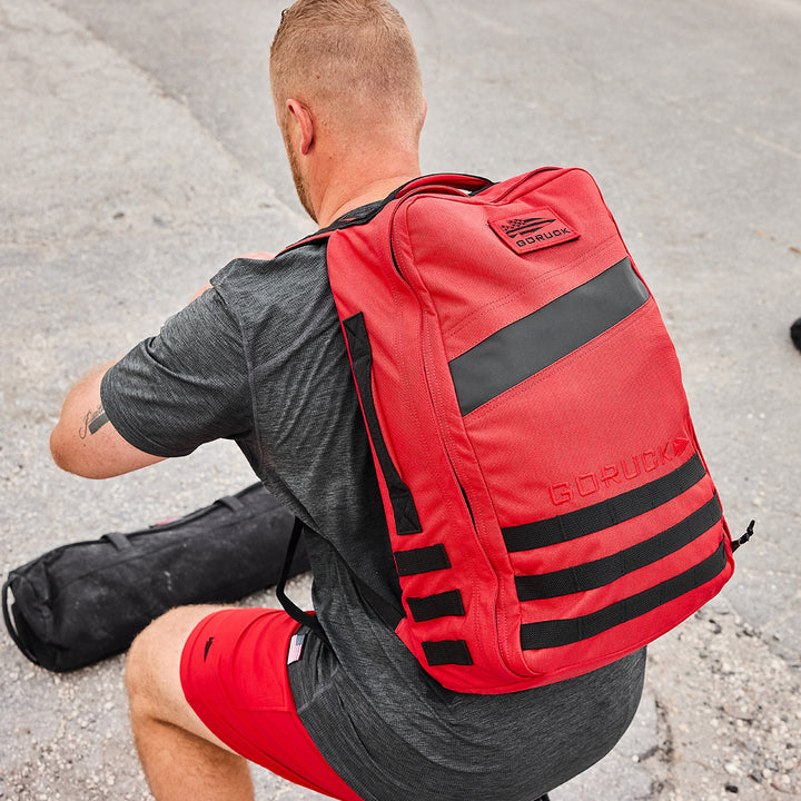 A man in a gray shirt and red shorts, sporting a red GORUCK Rucker 4.0 rucksack, is crouching on a concrete surface.