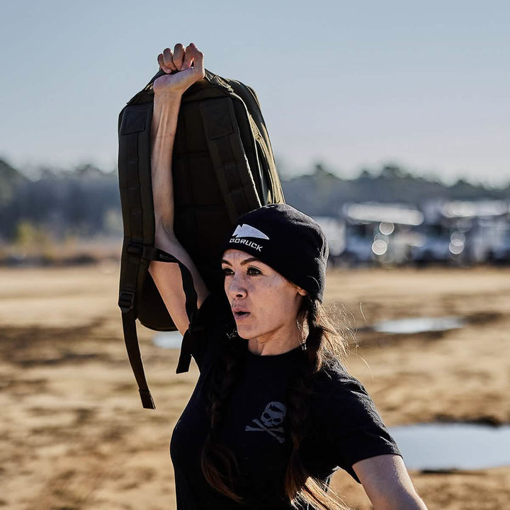 A person is energetically rucking outdoors, lifting a GORUCK Rucker 4.0 weighted backpack with one arm while wearing a beanie and black shirt, embracing the challenge with determination.