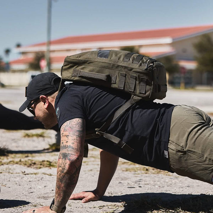 A person wearing GORUCK's Rucker 4.0 in casual outdoor gear is doing push-ups on the sand with a large green rucksack under a clear sky.