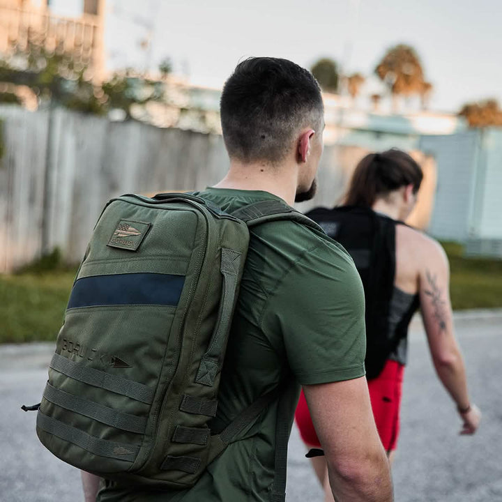 Two people enjoy rucking outdoors with GORUCK Rucker 4.0 backpacks—one in a green shirt and the other in a black tank top paired with bright red shorts.