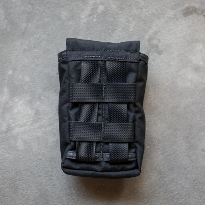 The GORUCK Simple Side Pocket, a black tactical pouch with straps, rests on a gray textured surface and includes MOLLE webbing and VELCRO® panels for versatile attachment.