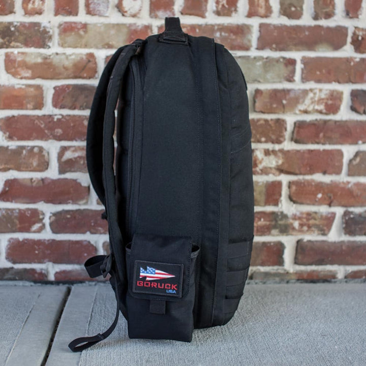 Simple Side Pocket by GORUCK, a black backpack with an American flag patch, displayed against a brick wall background and featuring MOLLE webbing for customizable storage.