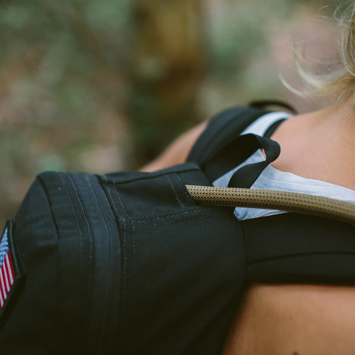 Close-up of a person wearing a backpack adorned with an American flag patch and a hydration tube, highlighting the Source Hydration Bladder, recommended by Special Forces for its built-in antimicrobial technology.