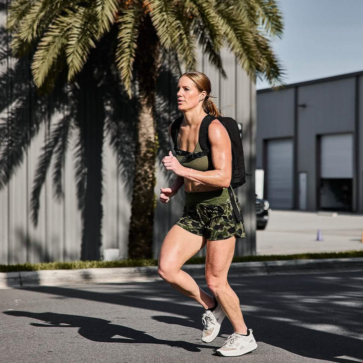 A person jogs outdoors in a sporty outfit with a backpack, featuring the breathable Stealth Bra - ToughFlex by GORUCK and green camouflage-style sports top and shorts. The background includes a palm tree and industrial buildings under a clear sky.