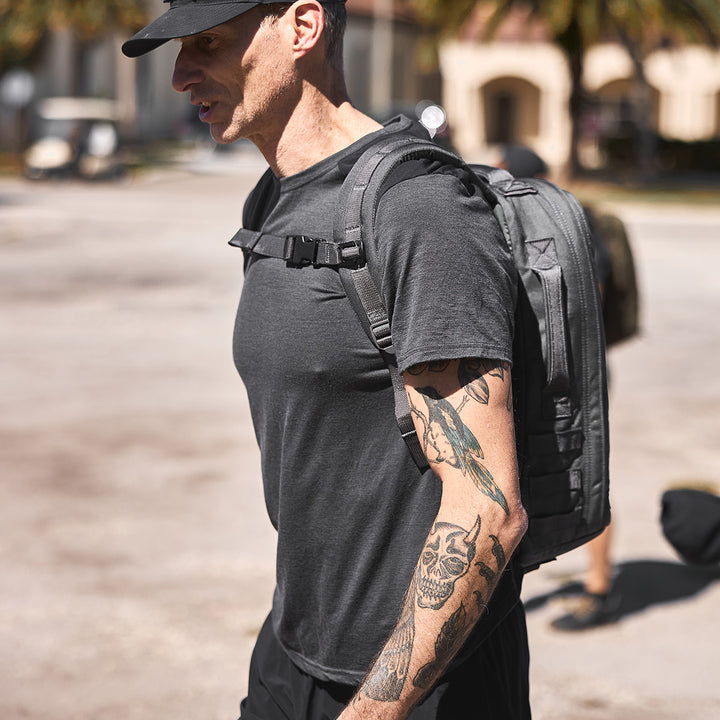 A tattooed man in a black shirt and hat confidently walks outside with a gray GORUCK backpack, enhanced by its MOLLE webbing for adjustable weight distribution and featuring the Training Sternum Strap for added comfort.