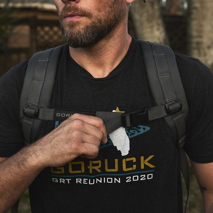 Man outdoors in a black "GORUCK GRT Reunion 2020" shirt adjusts a backpack strap with his left hand, ensuring optimal weight distribution with the Training Sternum Strap and MOLLE webbing.